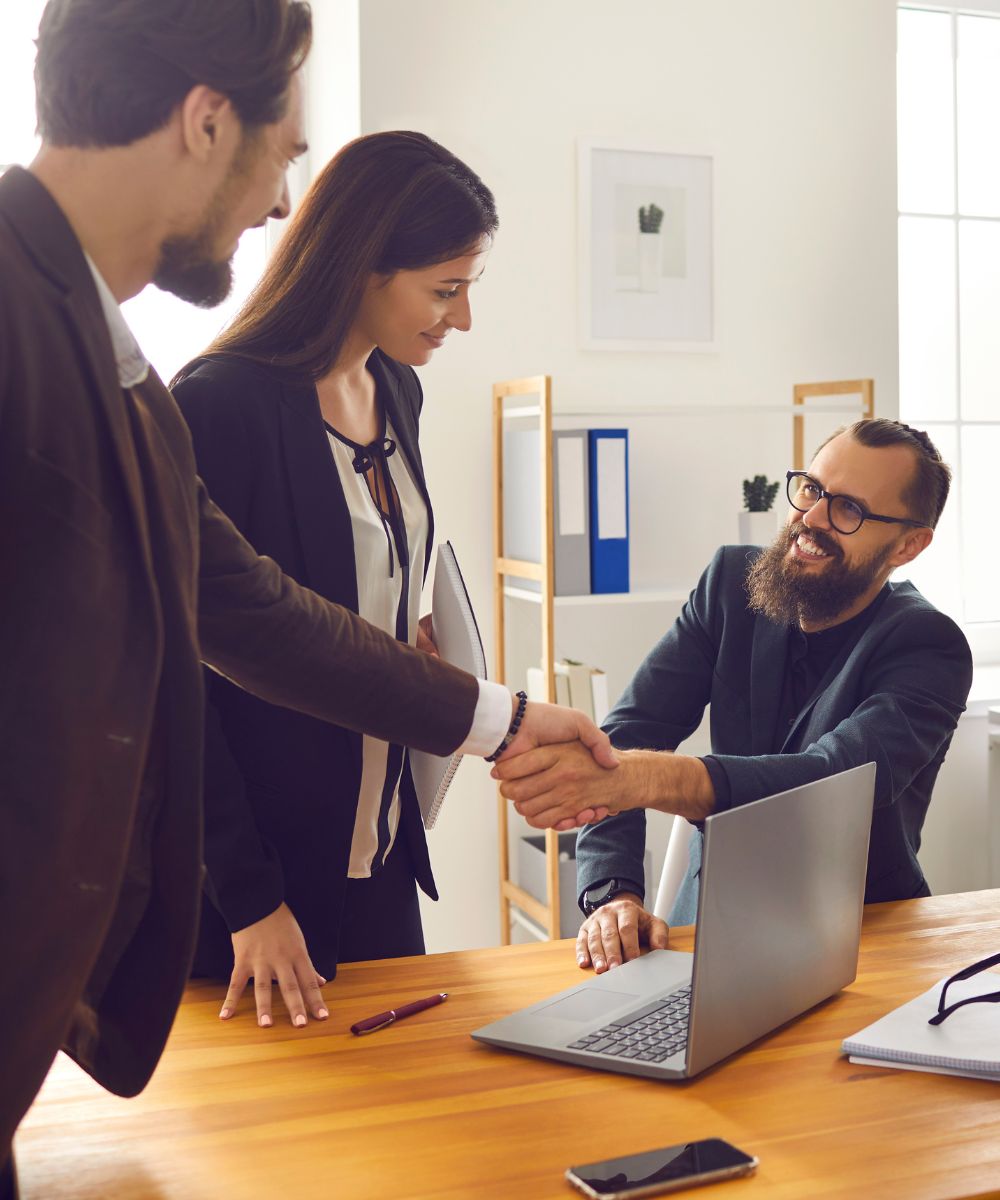 Person seated at desk shaking hands with a person standing while another person looks one Person seated at desk shaking hands with standing person as other standing person looks on.