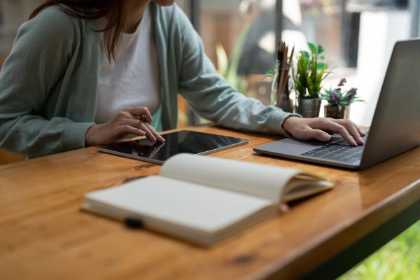 Cropped person seated at computer using laptop and tablet, open book on desk